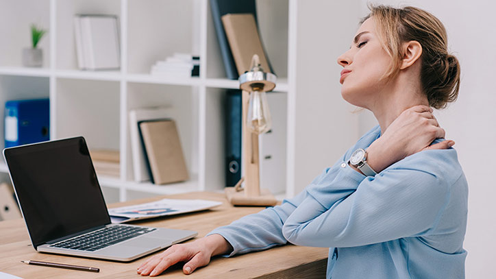 Eine Frau am Schreibtisch mit Laptop fasst sich mit geschlossenen Augen an den schmerzenden Nacken.