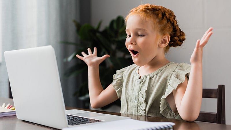 Ein kleines Mädchen sitzt vor einem Laptop und freut sich.