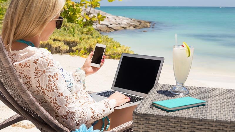 Eine Frau arbeitet mit Laptop und Smartphone an einem Strand, neben ihr ein Cocktail.