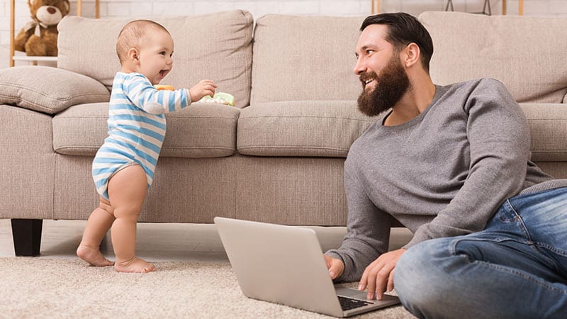 Ein Vater sitzt vor der Couch mit einem Laptop auf dem Boden. während neben ihm ein kleines Kind lacht.