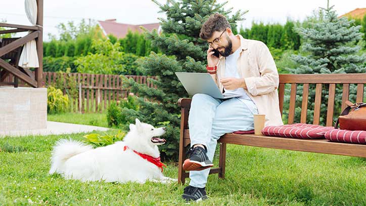 Ein Mann sitzt mit Laptop und Handy am Ohr auf einer Gartenbank im Grünen.