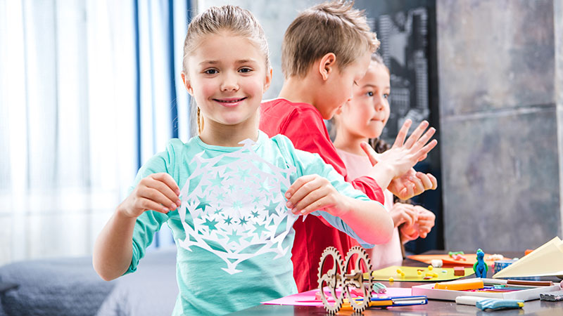 Drei Kinder basteln und ein Mädchen zeigt lachend ihr Ergebnis.