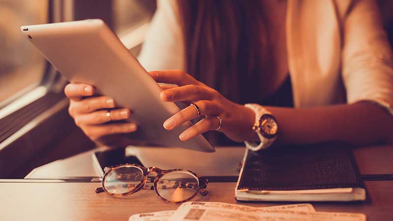 Eine Frau sitzt im Zug, in den Händen ein Tablet, vor sich auf dem Tisch ein Smartphone, ein Notitzbuch und eine Brille.