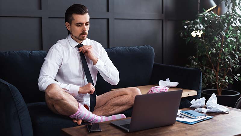 Ein Mann vor Laptop sitzt in Unterhose und mit Hemd und Krawatte auf der Couch.