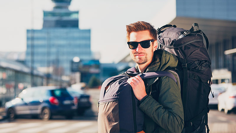 Ein Reisender mit Sonnenbrille und zwei großen Rucksäcken steht voller Vorfreude am Flughafen.