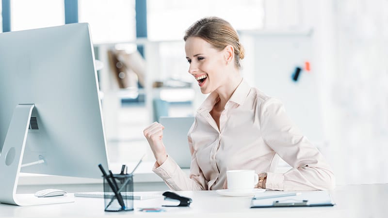 Eine Frau am Schreibtisch vor dem Monitor lacht und ballt die Faust.