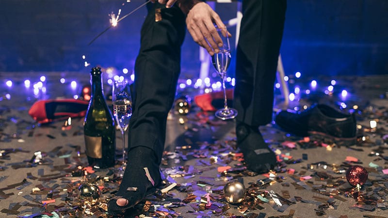 Ein Mann ohne Schuhe, mit zerissenen Socken und Glas in der Hand steht auf Resten von Weihnachtsdekoration