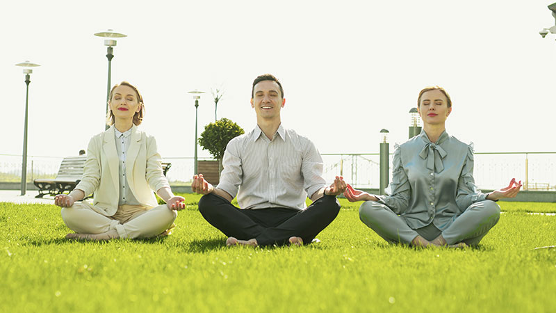 Drei Geschäftsleute sitzen nebeneinander im Schneidersitz auf der grünen Wiese und meditieren.