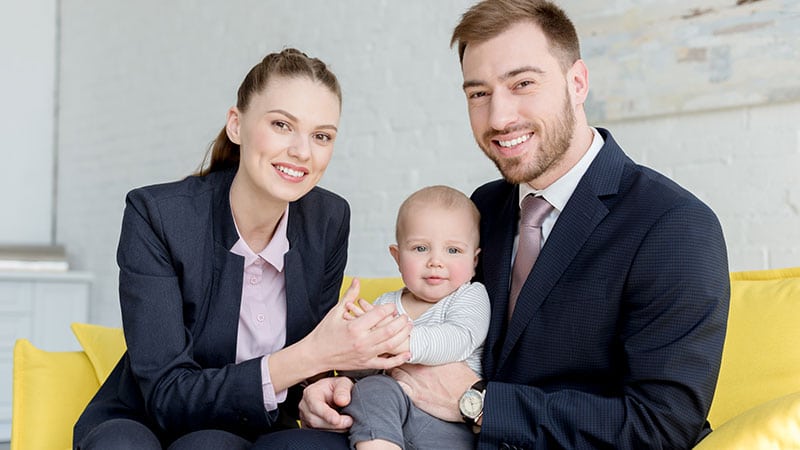 Ein junger Mann und eine junge Frau in schwarzen Geschäftzanzügen sitzen auf der gelben Couch mit einem Baby auf dem Schoß