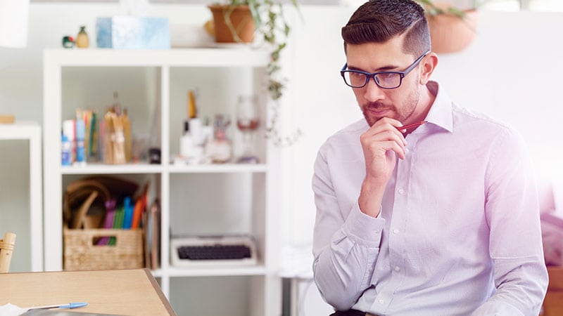 Ein junger Mann mit einer Brille sitzt nachdenklich in einer hellen Büroumgebung.