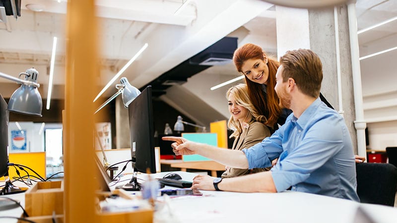 Zwei junge Frauen und ein junger Mann sitzen am Arbeitstisch.