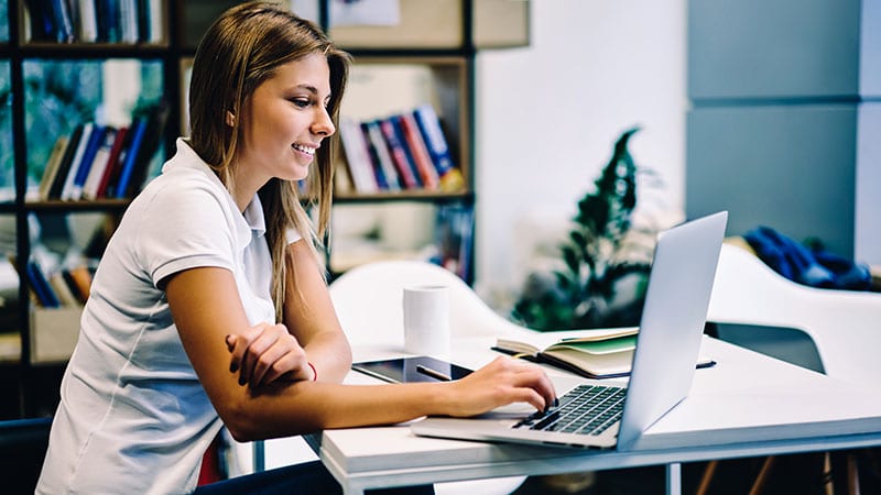 Eine junge Frau sitzt am Tisch mit einem Laptop und lächelt