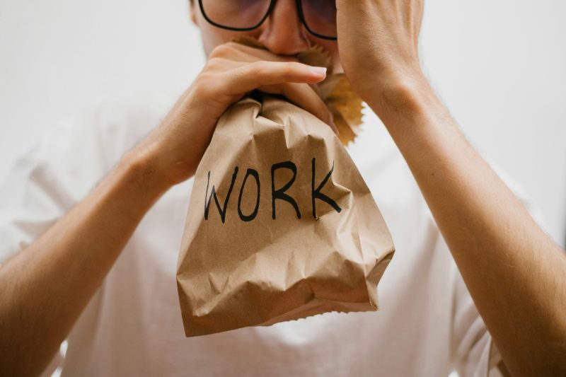 Person bläst in eine braune Papiertüte mit der Aufschrift „WORK“ — Symbol für beruflichen Druck