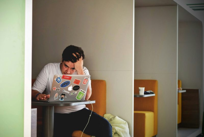 Person sitzt erschöpft an einem Tisch und stützt den Kopf, Laptop vor sich.
