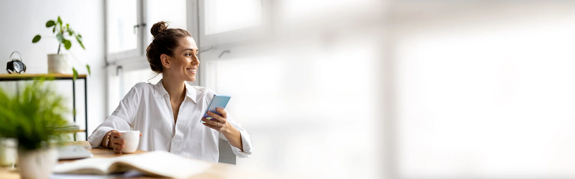 Junge Frau im Büro trinkt Kaffee und schaut auf ihr Smartphone.