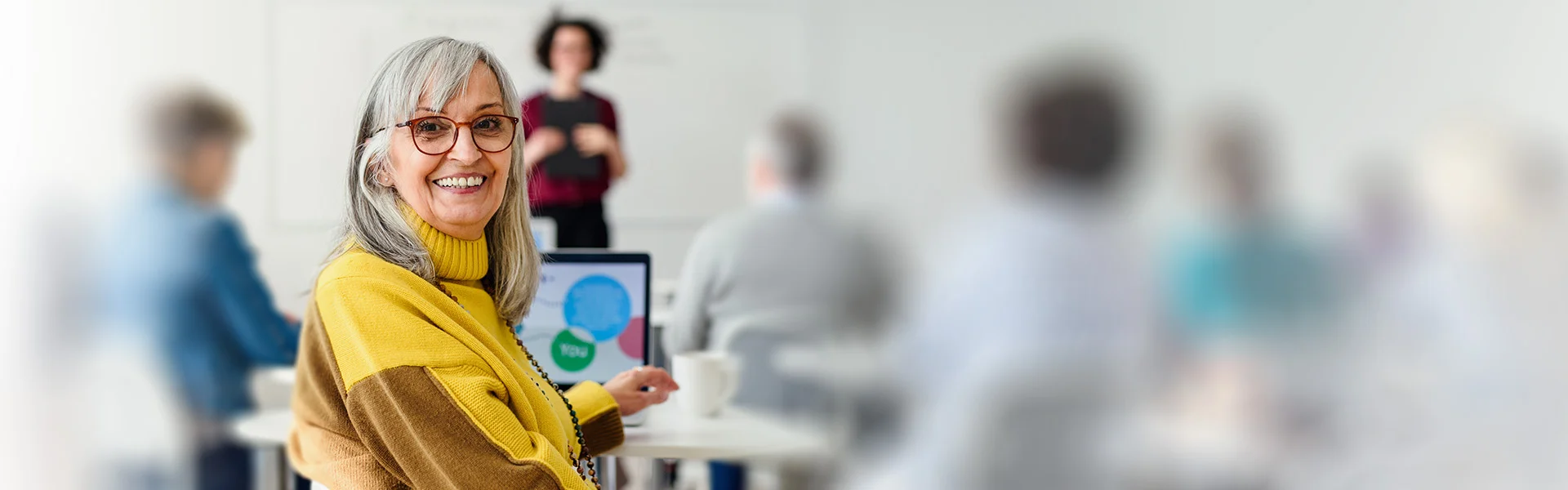 Frau mit langen grauen Haaren sitzt in einem Schulungsraum und nimmt am Laptop an einem Coaching für über 60-Jährige teil.