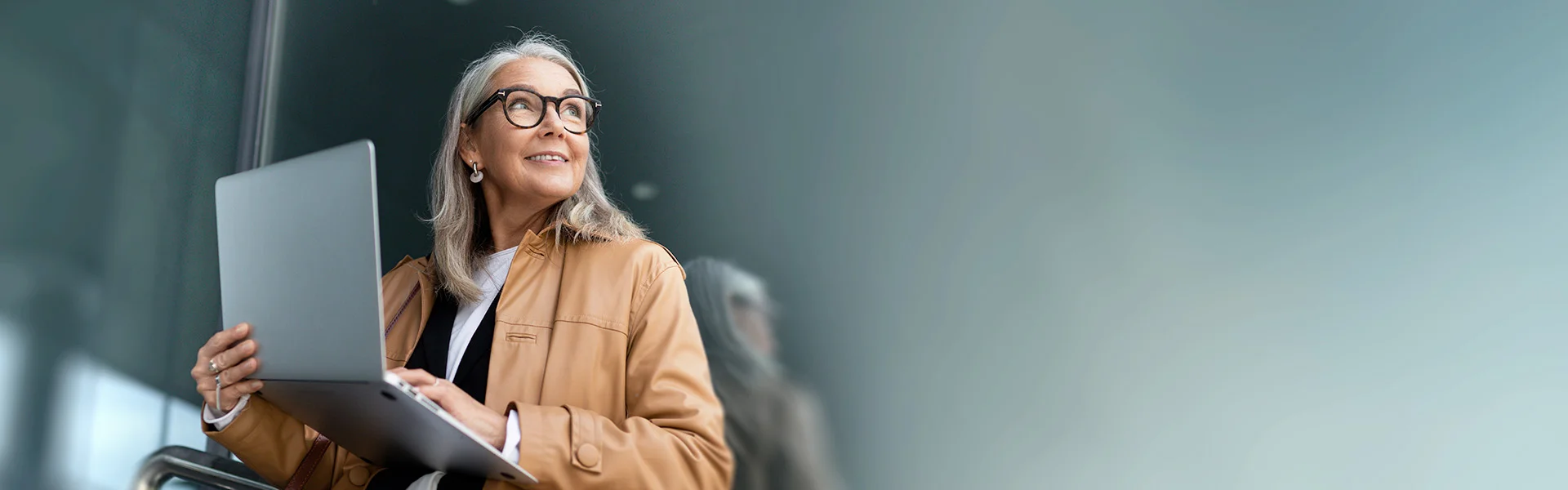 Eine Frau über 50 sitzt mit ihrem Laptop in beiden Händen und auf dem Schoß auf einem Stuhl und schaut zur Seite.