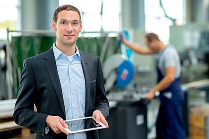Ein Mann in Anzug hält ein Tablet in der Hand, hinter ihm betätigt ein Arbeiter eine Maschine