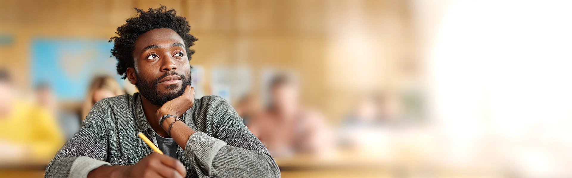 Ein dunkelhäutiger Mann sitzt im Klassenzimmer mit einem Stift in der Hand, das Kinn in der anderen Hand, und schaut nachdenklich zur Seite, während er über seiner Aufgabe brütet.