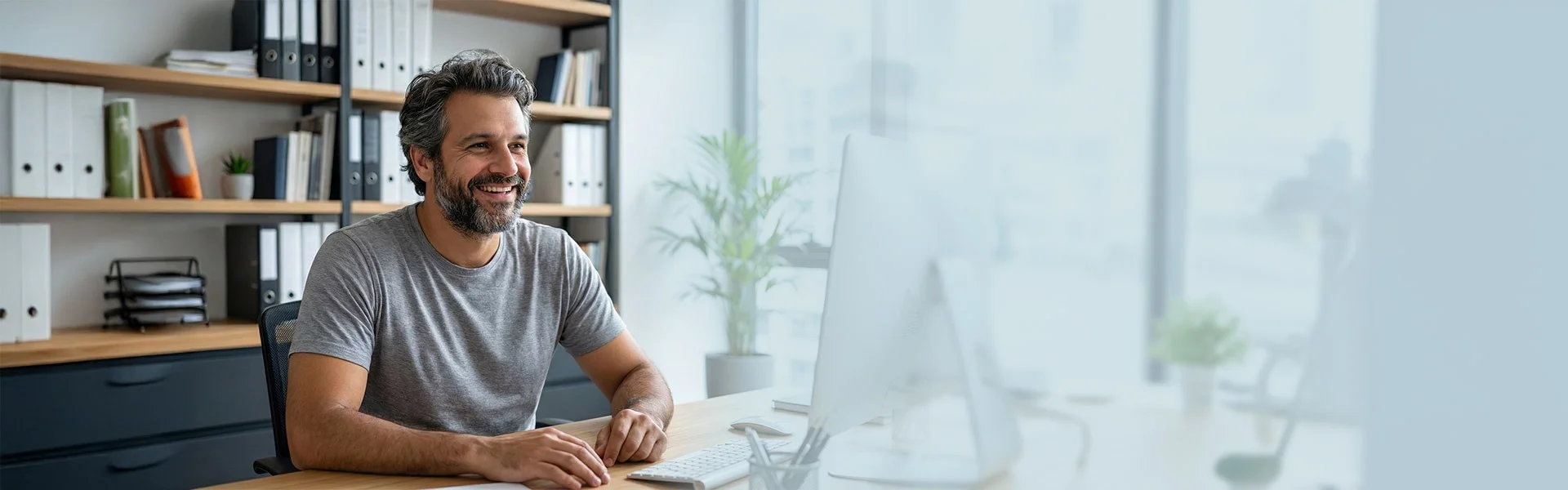 Ein bärtiger Mann sitzt vor einem PC und lächelt.