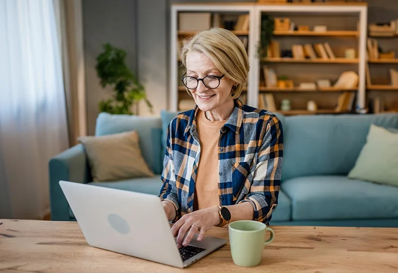 Frau in den 50ern sitzt im Wohnzimmer vor einem Laptop und lernt für ihre Weiterbildung.