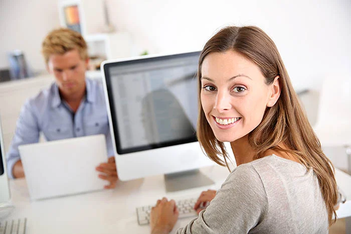 eine Frau, die an einem Schreibtisch mit einem Computer sitzt und mit einer anderen Frau spricht