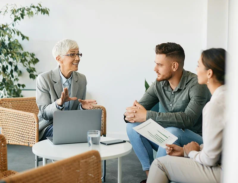 Eine Beraterin sitzt mit ihrem aufgeklappten Laptop zusammen mit ihren Kunden, einem Mann und einer Frau, um einen runden Tisch und erklärt etwas.
