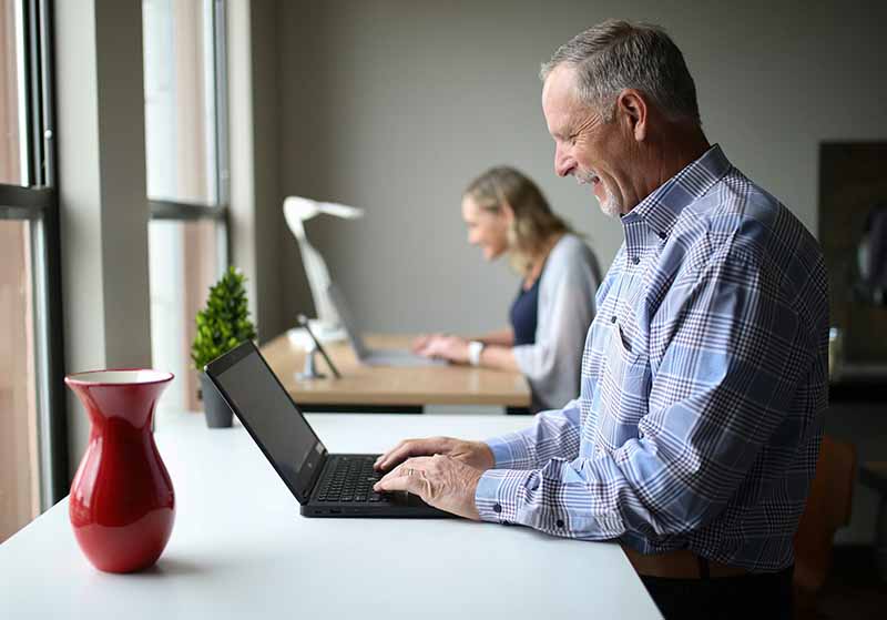 Vorn ein älterer Herr vor einem Fensterarbeitsplatz, im Hintergrund eine junge Frau am zweiten Schreibtisch.
