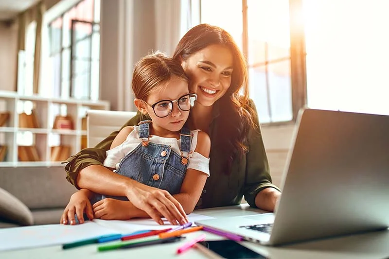 Eine junge Mutter sitzt zu Hause vor ihrem Laptop und arbeitet, während sie ihre Tochter, die sich an sie lehnt, im Arm hält.  