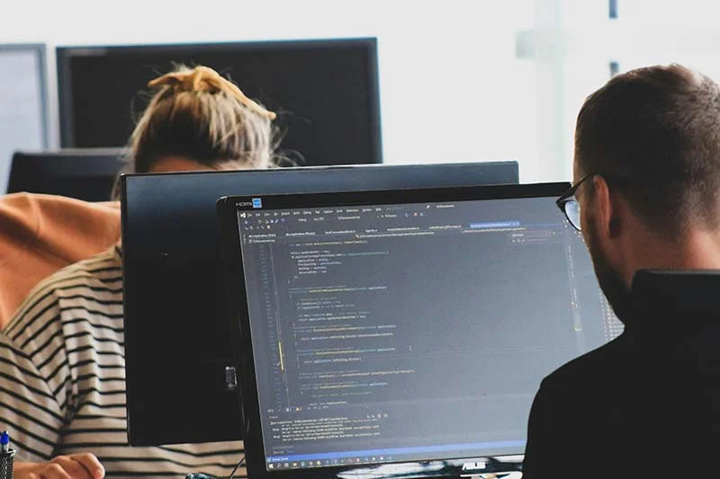 zwei Personen sitzen gegenüber an schreibtischen mit Bildschirmen in der Mitte. Es wird deutlich, dass sie was programmieren