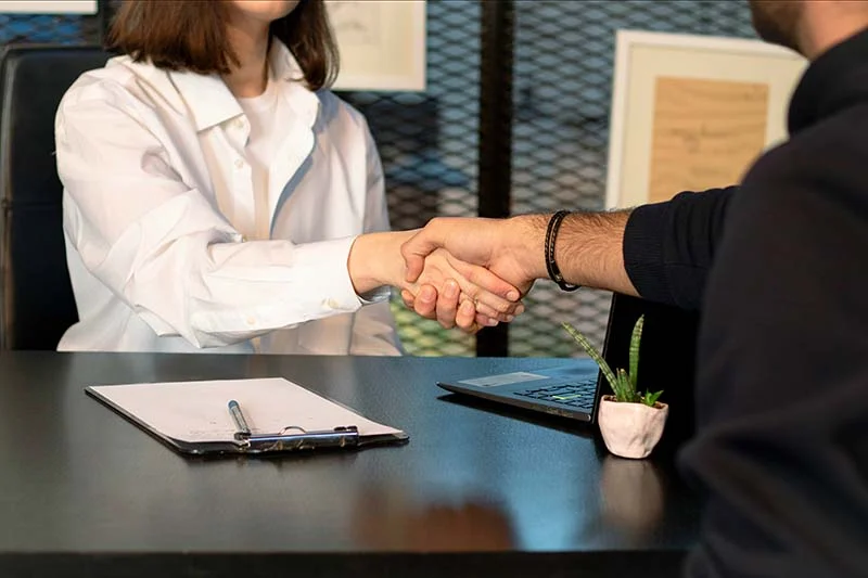 Eine Frau in weißer Bluse und ein Mann in schwarzem Shirt sind bis zum Kinn dabei zu sehen, wie sie sich über den Schreibtisch der Frau die Hand geben.