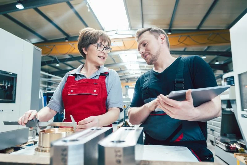 Symbolbild: Zwei Mitarbeitende besprechen konzentriert Messdaten an einer Werkbank; eine Person hält ein Tablet, die andere misst ein Bauteil.