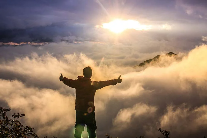 Mann steht mit ausgebreiteten Armen auf einem Berg und schaut über die Wolken.
