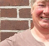 Die stolze Weiterbildungsteilnehmerin Astrid Estel steht, die Hände auf die Hüften gestützt, vor einer geziegelten Hauswand.