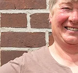 Die stolze Weiterbildungsteilnehmerin Astrid Estel steht, die Hände auf die Hüften gestützt, vor einer geziegelten Hauswand.