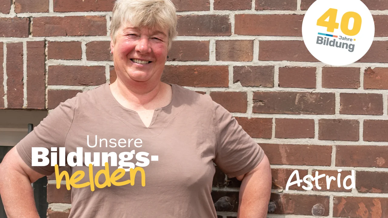 Video-Vorschaubild aus der Reihe "40 Jahre IBB - Bildungshelden": Die stolze Weiterbildungsteilnehmerin Astrid Estel steht, die Hände auf die Hüften gestützt, vor einer geziegelten Hauswand.