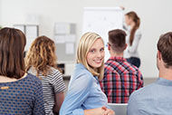 eine Gruppe von Menschen, die in einem Raum sitzen und ein Meeting haben. Eine blonde Frau dreht sich in Richtung Kamera