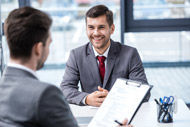 zwei Männer sitzen an einem Tisch und reden, einer hält eine Checkliste