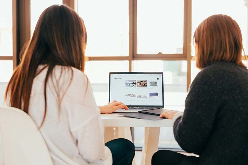 Blick von hinten auf zwei brünette Frauen, die am Tisch vor einem Laptop sitzen.