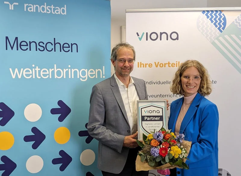 Dr. Christoph Kahlenberg (Manager Randstad Akademie & Arbeitsmarktprojekte) und Lea Tornow (Vorständin des IBB) stehen vor einem Randstad und einem Viona-Rollup und besiegeln mit einem Blumenstrauß und dem Viona-Partner-Zertifikat feierlich die Kooperation.